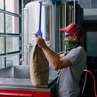 A man in a red baseball cap and a green bandana holding a big brown bag of cane sugar, a fermentation tank in the background.