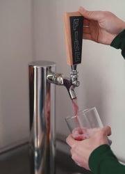 A closeup on two hands holding the tap and a glass and pouring Buchi Kombucha Fire.